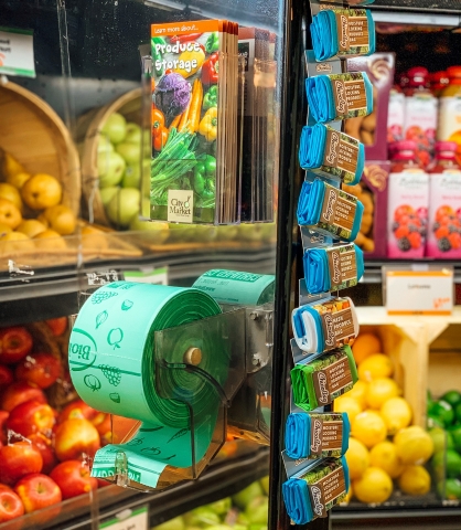 A roll of green compostable produce bags on a rack under a stack of brochures. The brochures have white text in front of a photo of assorted vegetables that reads "Produce Storage." Behind the rack is a produce display featuring brown baskets tipped on their sides. The baskets contain apples, pears, lemons and limes.