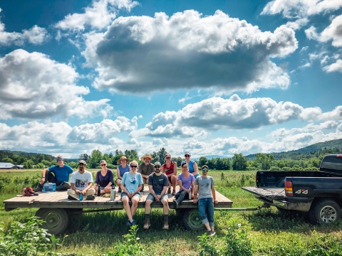 A group of City Market Members sitting on a flat trailer hitched to a green pickup truck. The truck is parked in a lush green field. The top half of the photo is rich blue sky dotted with fluffy clouds that are lit from above, with white tops and dark gray bottoms.