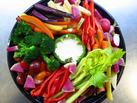 A round plate pictured from the top down. It is full of vegetables. Clockwise from top, there are thin slices of carrot that are orange, purple and white. There are thin slices of vividly red bell peppers. There are dark red cherry tomatoes. There are stalks of light green celery with fronds still attached. There are dark green broccoli florets. At center, a white dipping sauce is contained inside a green bell pepper.