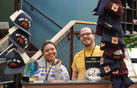 Two people stand behind a counter. The person to the left is wearing a gray sweatshirt and a lanyard. She has her left hand raised in a half-wave. The person to the right is wearing a yellow tee shirt and a lanyard. Both are smiling. To either side of the people, there are columns of hats. The hats on the left are trucker hats with the words "may all be fed" and pictures of carrots printed on them. The hats to the right are blue and burgundy beanies.