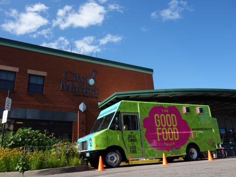 Green truck with "Good Food" logo on side in pink and yellow is parked in front of the downtown City Market Co-Op. The day is bright, the sky is blue, and there is a blooming garden next to the truck.