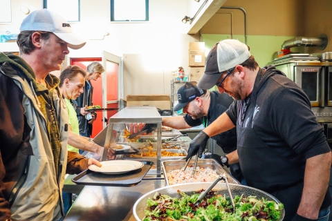 Two City Market staff members are serving food with tongs to three people holding trays with plates on them. There is a big salad bowl filled with leafy greens in the foreground.
