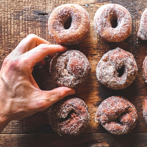 Six dark brown donuts are arrayed on a wooden table. They are dusted with white sugar. A hand is reaching for the left center donut.
