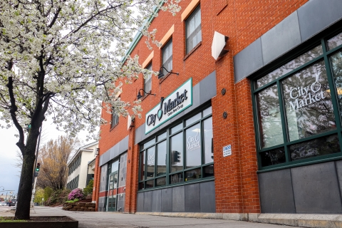 City Market Co-op downtown store exterior featuring brick building and a sign reading "City Market". There is a tree in the image in front of the store blooming with small white flowers. 