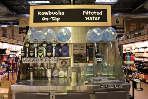 A large display with a slate gray sign with white lettering at the top. The left side of the sign reads "Kombucha on tap." Below that part of the sign are four brightly colored taps. The right side of the sign reads "Filtered Water." Below that part of the sign are two metal spouts. Below the taps and the spouts, there is a metal countertop.