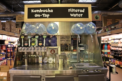 A large display with a slate gray sign with white lettering at the top. The left side of the sign reads "Kombucha on tap." Below that part of the sign are four brightly colored taps. The right side of the sign reads "Filtered Water." Below that part of the sign are two metal spouts. Below the taps and the spouts, there is a metal countertop.