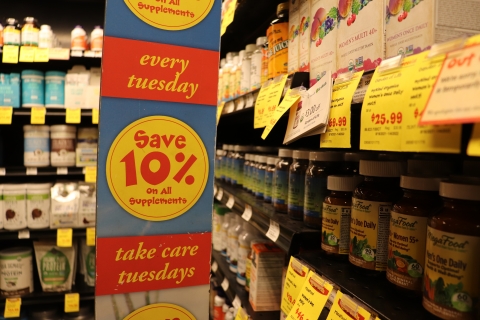 Across the left third of the picture, a vertical banner reads "Every Tuesday save 10% - Take care Tuesday." Behind the sign and slightly out of focus there is shelving containing a variety of small bottles with varied labels.