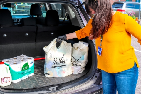 A person with long brown hair, a goldenrod shirt and jeans places City Market shopping bags into the back of a hatchback car, next to a red-capped gallon of milk and a twelve-roll pack of toilet paper.