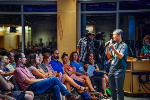 People seated in two rows in a large room. Behind them are tall windows. Outside the windows, it's dark. In front of the windows, a man in a checked shirt holds a video camera. At the far right of the image, a well-lit man with short black hair holds a microphone. His free hand is raised in an expressive gesture. He is wearing jeans and a blue-gray tee shirt.