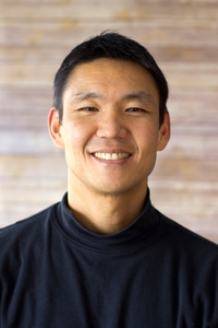 A portrait of John Tashiro wearing a black turtleneck and sitting in front of a rustic wooden background