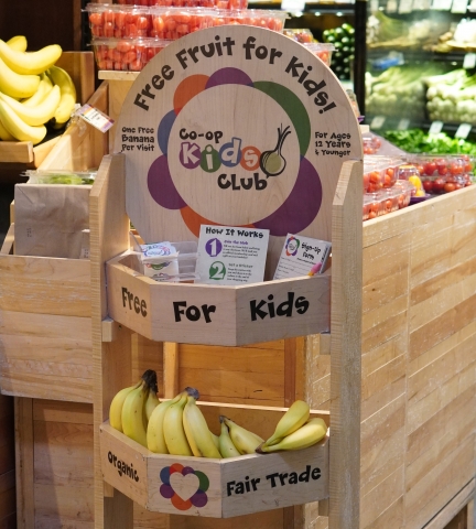A display made of light brown wood with a round sign on top. The sight reads "Free fruit for kids! Co-op kids club." In the first shelf of the display there are white brochures with black writing and purple and green graphics. In the second shelf, there is an assortment of ripe yellow bananas.