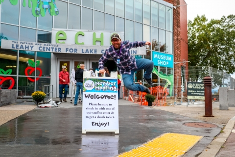 At the center of the image, a rectangular sign reads "Welcome!" Above the main text on the sign is a City Market logo and the words "Member Meeting." A man in a green baseball cap, a blue and white plaid shirt and jeans is holding onto the top of the sign and doing a frog kick. The man and the sign are on the sidewalk in front of a glass-fronted building with a sign that reads "echo."