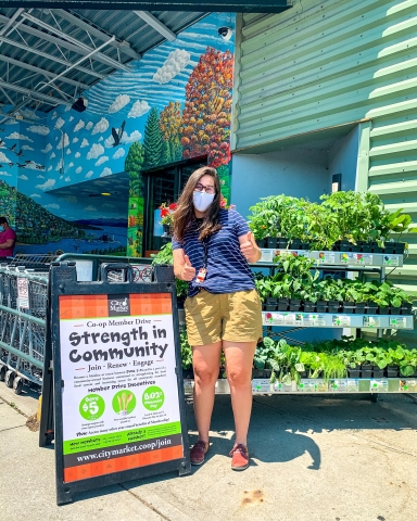 A woman stands to the right of a waist-high sign that reads "Strength In Community." She's wearing a blue tee shirt and yellow shorts, and she's giving two thumbs up. The woman and sign are in front of a rack of potted plants, and the rack of plants is in front of City Market's downtown entrance.