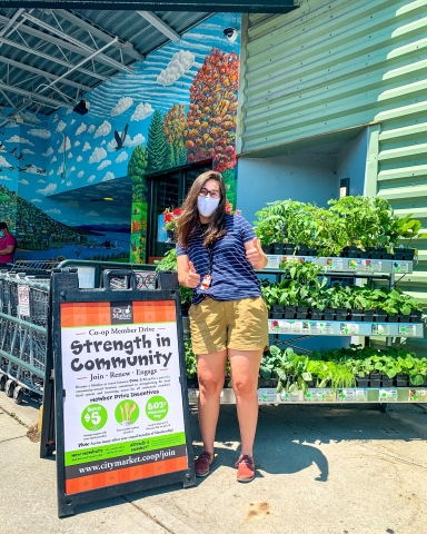A woman stands to the right of a waist-high sign that reads "Strength In Community." She's wearing a blue tee shirt and yellow shorts, and she's giving two thumbs up. The woman and sign are in front of a rack of potted plants, and the rack of plants is in front of City Market's downtown entrance.