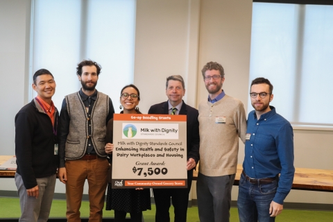 Six people stand in a row in a white-walled conference room. The two people in the middle are holding a sign that has "Milk With Dignity" printed at the top and "$7,500" printed at the bottom.