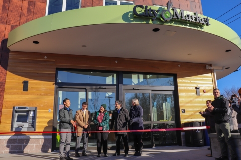 Five people stand in front of a building with sliding doors and a marquee that reads "City Market." There is a red ribbon in front of the people and all along the entrance to the building. The person to the farthest left is talking, and the other four face him. At the far right, several additional people are standing and clappng.