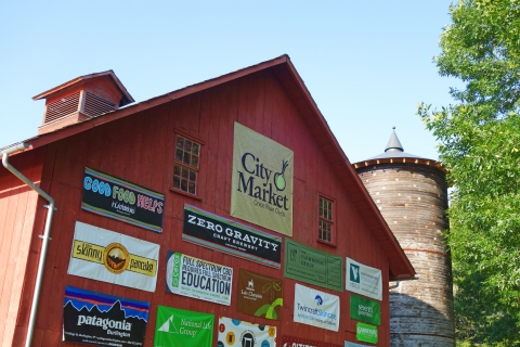A red barn with large banners featuring logos of different companies and organizations. At the top, there is a brown square banner with "City Market" printed on it in black lettering. There is an abstract illustration of an onion in the upper right of the banner.