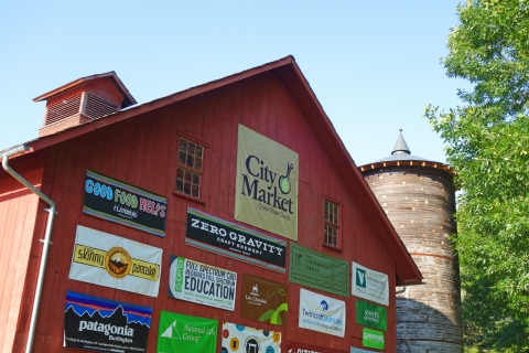 A red barn with large banners featuring logos of different companies and organizations. At the top, there is a brown square banner with "City Market" printed on it in black lettering. There is an abstract illustration of an onion in the upper right of the banner.