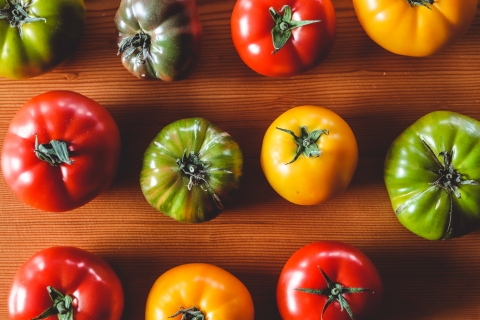 Three rows of heirloom tomatoes on a wooden table, viewed from the top down. The tomatoes are lush, brightly lit and in a variety of colors, including red, yellow, light green, and mottled red and dark green.