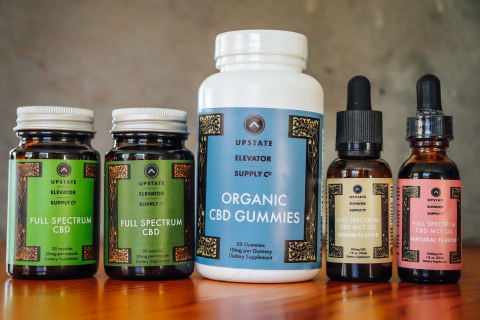A row of Upstate Elevator Supply Company CBD products on a shiny wooden table in front of a mottled gray wall. The items are two green-labeled jars, one large blue-labeled jars, and two glass bottles with droppers, labeled in yellow and pink.