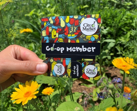 A hand holding a Co-op Membership card. The card depicts brightly-colored abstract art with a City Market logo in the upper right. Across the bottom, it reads "Co-op Member" in white letters. The card is being held in front of a wildflower garden.