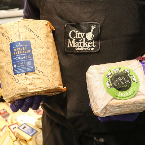 A person in a black apron with the City Market logo in the middle of it holds two paper-wrapped bundles of cheese. The one on the left is labeled "Bayley Hazen Blue." The one on the right is labeled "Orb Weaver."