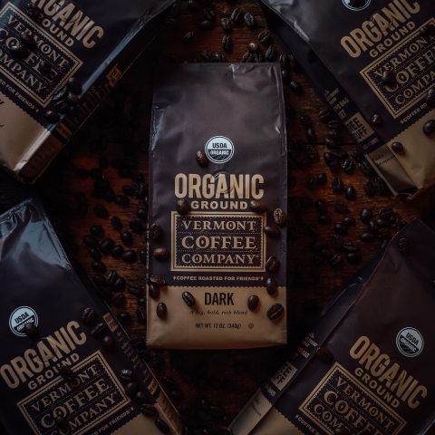 A dark brown bag at center labeled "Organic ground VT Coffee Company Dark." The bag is face up on a bed of coffee beans, which are very dark brown.