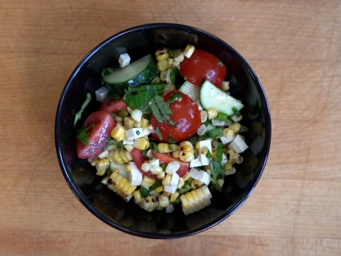 A top-down view of a black bowl with a completed grilled corn and zucchini salad with vibrant red cherry tomatoes, white ricotta salata cheese, and green chopped basil against a wooden cutting board.