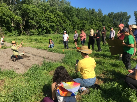 12 people are arranged in a semicircle at the right of the photo. Some are sitting and some are standing on a grassy field. To their left, a person is sitting in a field of dirt. They are wearing a green shirt and brown hat, and they are holding a sapling.