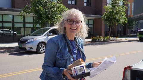 A woman with curly white hair and black rimmed glasses stands in front of a street. She is wearing a blue denim jacket, holding a few sheets of paper, and smiling widely.