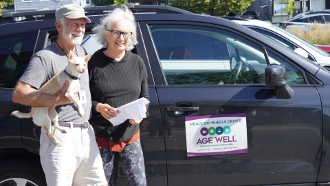 A man with a tan hat, grey t-shirt, white beard, and khaki shorts stands with a woman with white shoulder-length hair, black sweatshirt, and patterned leggings. The man holds a Chihuahua with his right arm, and his left arm is around the woman. They are standing in front of a Black SUV with the Age Well logo on the passenger door.