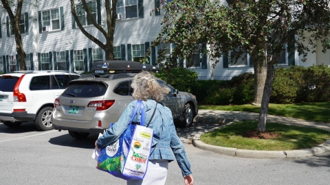 Woman with grey curly shoulder-length hair, wearing a denim jacket, and a City Market reusable grocery bag with her back facing the camera walks through a parking lot of white condo complexs with green shutters. There is a gold subaru outback and a white Volvo SUV in the parking lot. 
