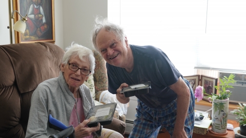 Man with white hair, grey t-shirt, and plaid pajama pants stands crouched over next to a woman sitting down with white short hair wearing glasses, a pink pajama top, and a grey zip-up on. They are both smiling and holding up their pre-packaged Age Well meals in a living room with white walls.