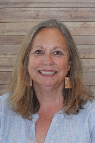 A photo of Trish Redalieu smiling wearing a light blue blouse with some white faint lines on it. She also is wearing tan square dangly earrings. She is sitting posed against a light wood background.