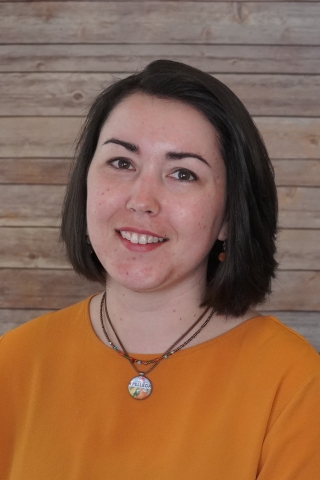 Claire Ross smiling in an orange sweater and two necklaces, one having a San Pellegrino cap on it. She is sitting against a light wood backdrop.