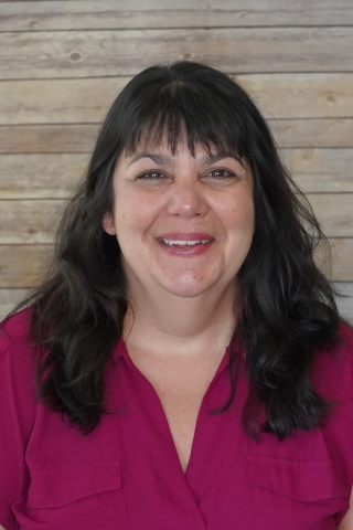 A photo of Madelyn Cataford smiling wearing a pink/maroon blouse with pockets. She is seated against a light wood background.