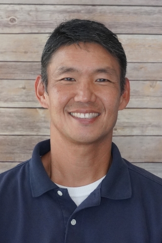 A photo of John Tashiro wearing a navy blue polo with white buttons and a white t-shirt underneath showing near the top. He is sittting down and smiling against a light wood back drop