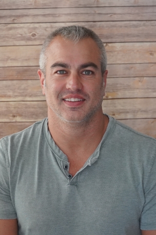 A photo of Brent Demers smiling wearing a light grey-blue henley short sleeve t-shirt with two buttons at the top. He is sitting against a light wood background.