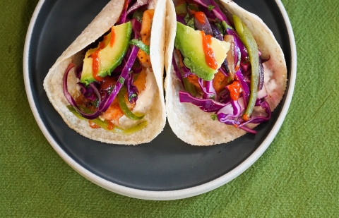 Two fish tacos on a slate gray plate. The tacos are topped with green avocado, purple cabbage slaw and orange hot sauce.