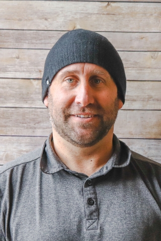 Matt Butterly pictured wearing black beanie and grey polo with black buttons. He is smiling against a light wood backdrop.