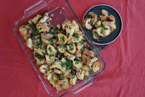 Chopped baguette, melted cheese, and chopped bacon topped with parsley in rectangle glass baking dish with right corner of the strata taken out. The remainder of the strata is on a grey plate to the right of the baking dish, with both the plate and the dish presented on a red table cloth. 