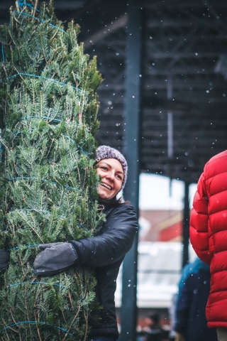 A person wearing a black coat and a knit hat hugs a tree. The person is smiling. Around the person and the tree, white heavy snowflakes drift in the air.