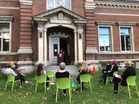 A person in a red coat stands at a podium in front of the Fletcher Free Library speaking through a microphone to people in green chairs.