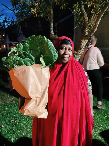 A woman with a soft smile on wearing a red garment on her head that spans the length of her body. She is holding a brown paper bag with leafy greens that are fanning out of the bag.