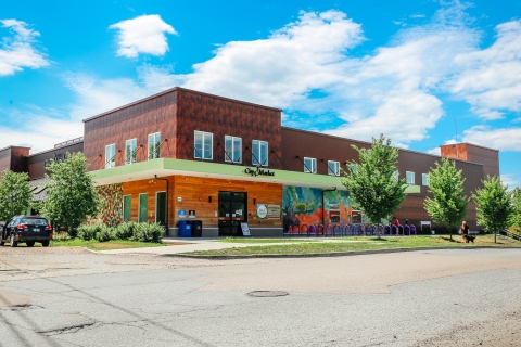The City Market South End Store as seen from Flynn Avenue. The store is reddish brown with a green overhang with the words "City Market" on it. At the right side of the door, there is a mural of a moose. In front of the moose, there are purple bicycle racks. The sky is bright blue with white puffy clouds.