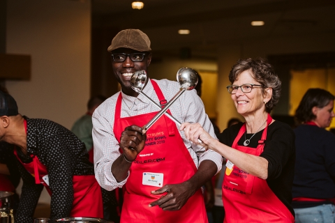 Ali Dieng and Rita Markley, both smiling, and holding two ladles across each other while wearing red aprons. The aprons read Spectrum's Empty Bowl Dinner 2019, and they are both wearing nametags. 