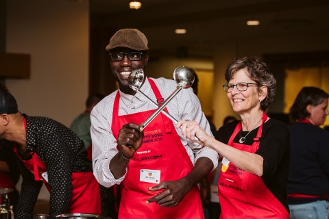 Ali Dieng and Rita Markley, both smiling, and holding two ladles across each other while wearing red aprons. The aprons read Spectrum's Empty Bowl Dinner 2019, and they are both wearing nametags.