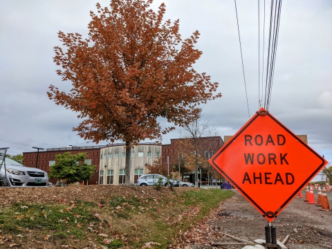 A bright orange diamond-shaped sign reads "Road Work Ahead." Behind it, a tree with a full, dusty orange crown stands in front of the City Market store in the South End. The sky is grey-blue.