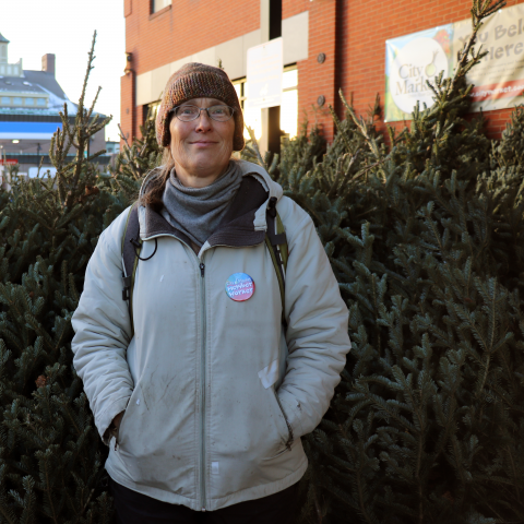 A person in a beige coat with a gray scarf tucked into it and a brown knit cap stands in front of a row of cut balsam trees. The person has a button pinned to their coat that has the words "Member Worker" printed on it in white puffy text.