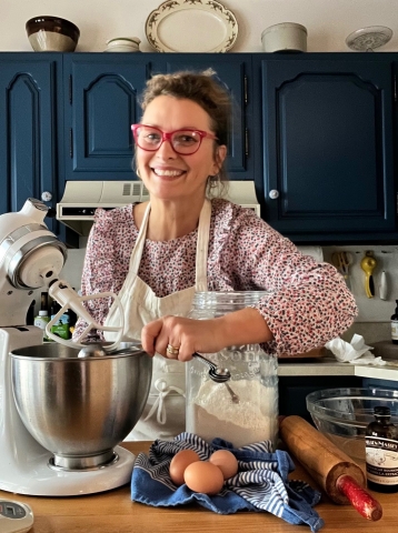 A woman wearing red glasses stands near a mixer and baking ingredients in a kitchen with blue cabinets